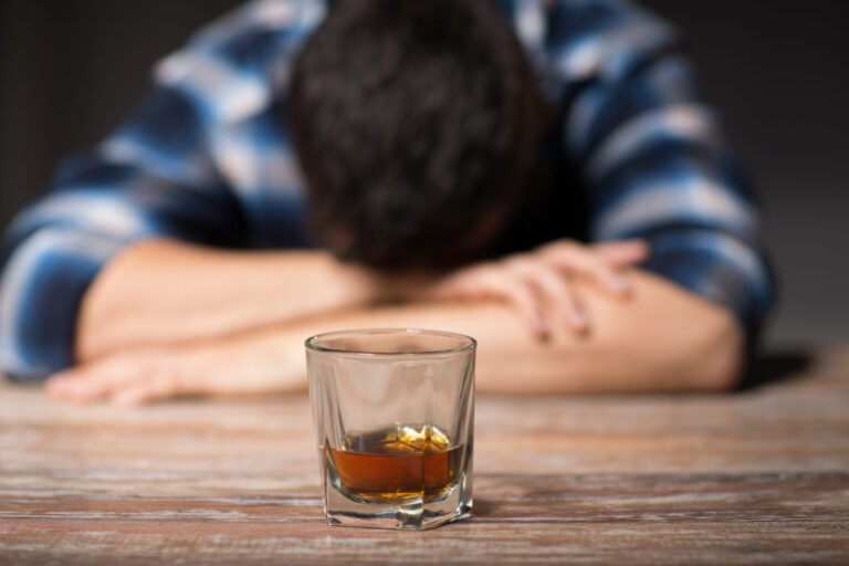 A man rests his head on his arms at a wooden table, a glass of whiskey in front of him