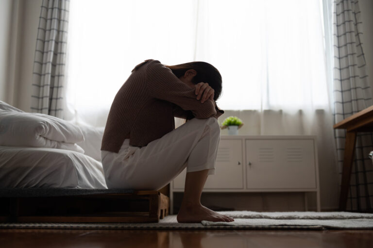 A woman sits hunched on the edge of a bed with her head buried in her arms